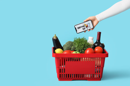 Female hand holding mobile phone with text SHOPPING DAY and red basket with different fresh products on blue background - Powered by Adobe