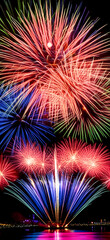 A spectacular display of colorful red, blue, and white fireworks bursting in the dark night sky over water, creating beautiful reflections.