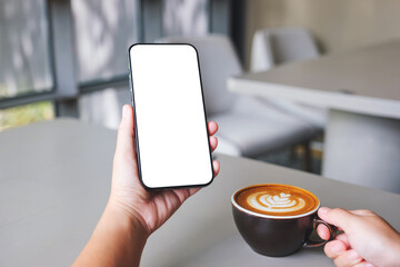 Mockup image of a woman holding and using mobile phone with blank desktop screen while drinking coffee in cafe