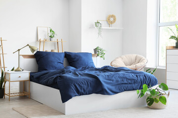 Bed with stylish blue bedding, armchair, shelving unit, bedside table and houseplants near window...