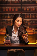 Woman in a black suit sits holding a glass of whiskey at a bar table.