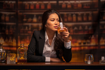 Woman in a black suit sits holding a glass of whiskey at a bar table.