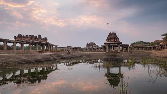 Time lapse: ancient stone temple complex in Hampi, India, reflected in water tank under dramatic evening sky, symbolizing heritage, spirituality, and timeless history