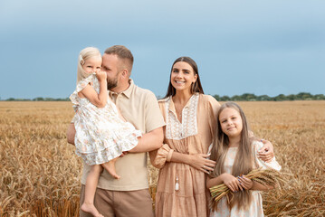 Family of four, two daughters, standing together in a golden wheat field, smiling joyfully, with a...