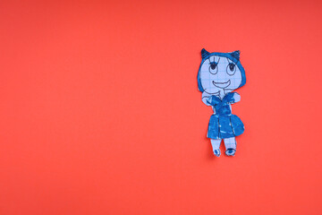 A small paper doll, cut out from a drawing, in a blue outfit and with ears, stands on a bright red background. The photograph demonstrates the simplicity of children's creativity and has copy space fo