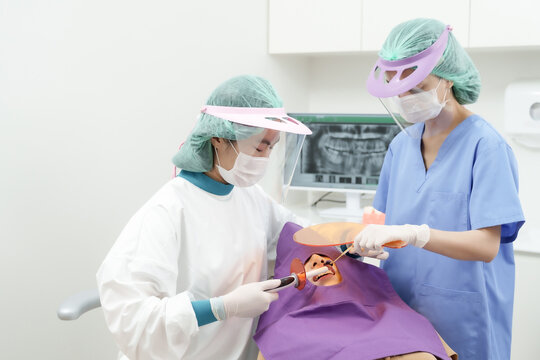 Asian adult woman in dental chair treated with curing light during composite filling, assistant applies light while dentist uses dental instrument, both professionals wear face shields