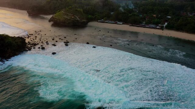 A breathtaking sunset over Watu Karung Beach, Pacitan, Indonesia. Golden light reflects on the ocean as waves crash around a large rock island. The vibrant scene captures the beauty of the coastal