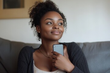 Happy African American woman sitting on a sofa at home, holding a phone to her chest with a dreamy expression, looking to the side with joy after receiving good news via text, Generative AI