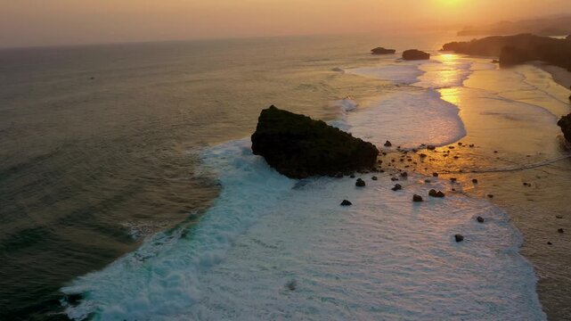 sunset over Watu Karung Beach, Pacitan, Indonesia. Golden light reflects on the ocean as waves crash around a large rock island. The vibrant scene captures the beauty of the coastal landscape.