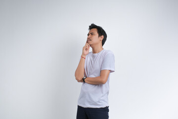 A young man in a white t-shirt standing with a thoughtful expression, hand on chin, and looking...