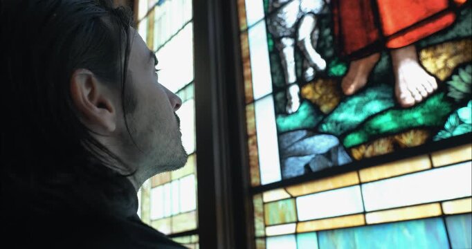 Closeup of man stands inside a church, gazing at a stained-glass window of Jesus the Good Shepherd, evoking themes of spirituality, faith, and quiet reflection.