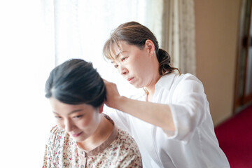 A woman with dark hair is getting her hair styled by a woman with lighter hair. They are in a room with a window