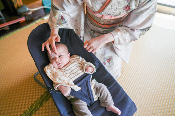 A woman is holding a baby in a blue high chair. The baby is wearing a striped shirt and has a white...