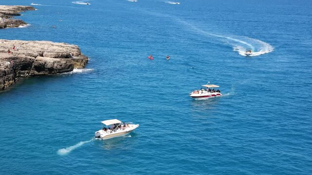 Sunny summer day in Polignano a Mare, Italy. Tourist boats float in the blue sea near crowded beaches and the historic stone bridge. Old buildings, cathedrals, and bustling streets. Mixed video
