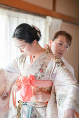 A woman is getting her hair styled by another woman, who is wearing a kimono. The woman getting her hair done is adjusting her kimono, possibly to make it fit better or to fix a loose piece