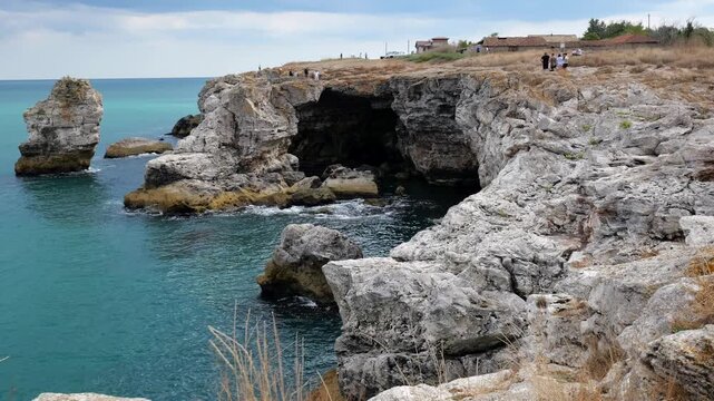 Sunny day at Tyulenovo, a famous tourist destination on the Black Sea near Shabla and Kavarna. Scenic rocky arch, caves, and sharp sea cliffs. Tourists walking along the cliffs and people jumping into