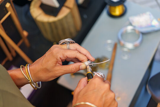 Metalsmith bending aluminum wire, creating intricate handcrafted jewelry beside colorful market display - Powered by Adobe