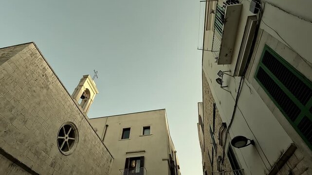 Chiesa di S. Andrea, Bari, Italy. Cinematic tilt shot from top to bottom showing old buildings, a narrow street with stone pavement, a cross, and the bell tower.
