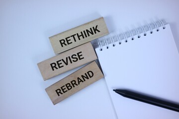 Obraz premium Wooden blocks with motivational words 'Rethink,' 'Revise,' 'Rebrand' displayed beside a blank notebook with pen on a vibrant white background