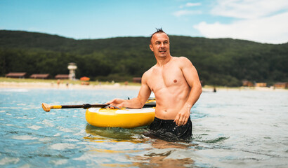 Fit man confidently holding paddle board in shallow water, looking into distance. Wet hair, muscular body, relaxed face pure masculine energy and enjoyment of the moment.
