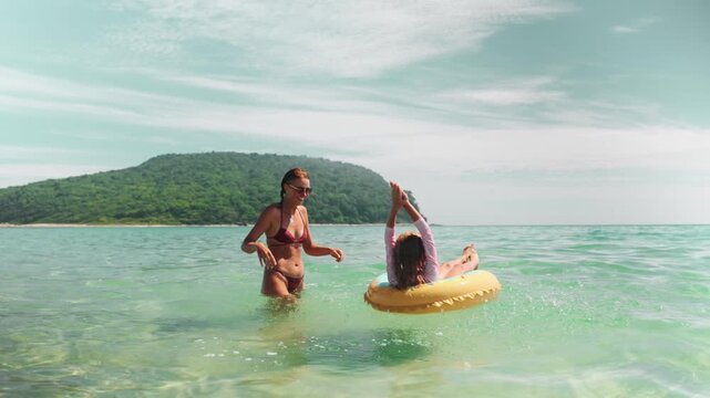  Joyful mom throws water as daughter laughs in inflatable ring. Beautiful moment of parenting, positive emotion and family interaction on beach vacation in crystal clear sea.