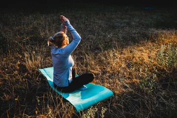 Fotobehang Lotusbloem Woman practicing yoga and meditation on nature sitting in lotus pose on yoga mat, relaxed with closed eyes. Mindful meditation concept. Wellbeing. Sunset meditation. Place for text.  © Antonina Polezhayeva