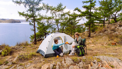 Friends enjoy tea beside tent near peaceful lake during sunny afternoon
