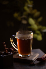 Hot tea is shown in a clear glass mug, with steam rising from the surface. The moody, dark...
