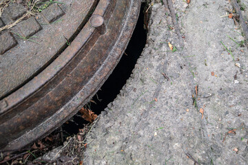 Detailed view of an old rusty manhole cover misaligned with concrete, showing a hazardous gap leading into darkness