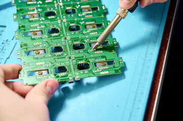 Hands assembling green circuit boards with a soldering iron in a workshop during daylight hours