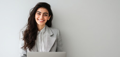 The woman in a gray blazer smiling at her laptop against a neutral wall