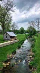 Obraz premium Tranquil landscape featuring a small cabin nestled beside a gently flowing stream under a cloudy sky, surrounded by lush green grass and trees