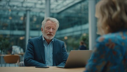 Senior man talks to woman, laptop on table. Indoor, bright, glass wall background