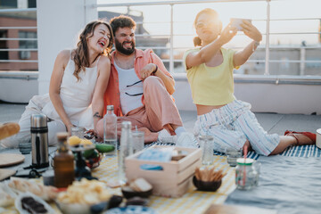Diverse friends having fun on a rooftop terrace, sharing laughter and joy during a sunset picnic while capturing moments with a selfie.