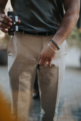 A close-up of a man dressed in light pants and a dark shirt, wearing bracelets, and holding a black device in an outdoor setting with a blurred background.