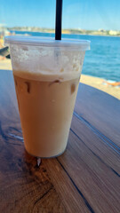 Iced coffee in a clear cup on a wooden table by the water. The background features a blue sky and calm water, creating a refreshing atmosphere.