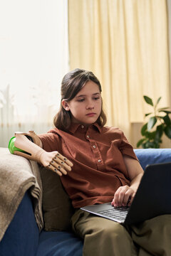 Teenage kid with prosthetic arm using laptop at home on sofa
