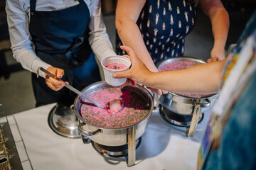 A close-up of people serving and receiving cold pink beet soup, likely traditional Latvian aukstā zupa, at a food service station. Suitable for commercial use.
