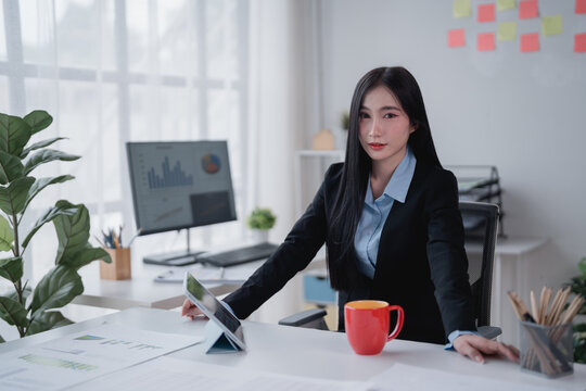 Young asian businesswoman sitting at desk using tablet and computer analyzing financial charts and graphs, working on business project in modern office with sticky notes on wall and plants