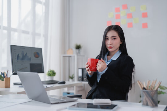 Young asian businesswoman drinking coffee during break in modern office with laptop, computer and sticky notes on background, business and finance work concept - Powered by Adobe