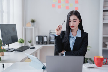 Young Asian businesswoman holding a pen and contemplating her work while using a laptop in a...