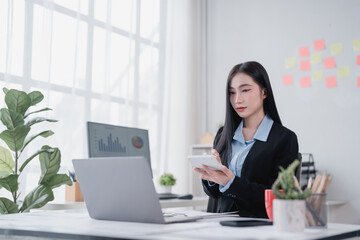Young Asian businesswoman using calculator and working on laptop computer with graphs on screen in...
