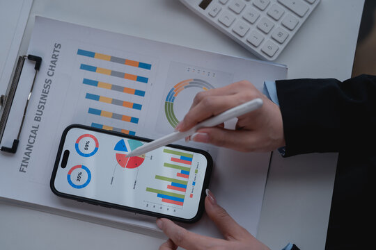 Businesswoman analyzing financial business charts displayed on smartphone screen using stylus pen. With documents and calculator on desk. Performing financial analysis and business strategy planning