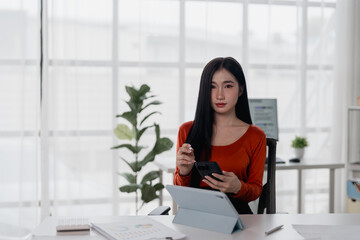 Young Asian businesswoman holding a smartphone and working on a tablet while seated at a desk in a...