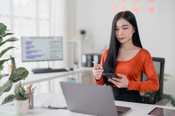 Young asian businesswoman using smartphone and stylus pen while working with laptop computer at...