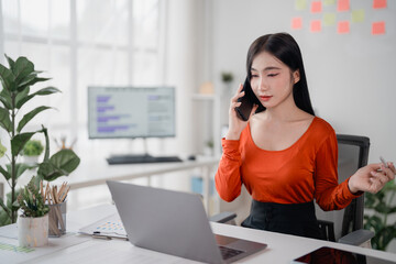 Young Asian businesswoman engaged in a phone conversation while working on her laptop in a modern office, skillfully managing her business operations from a distance