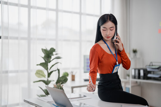 Young Asian businesswoman wearing an id card, talking on phone and taking notes while sitting on desk in modern office, she is working with laptop and documents - Powered by Adobe