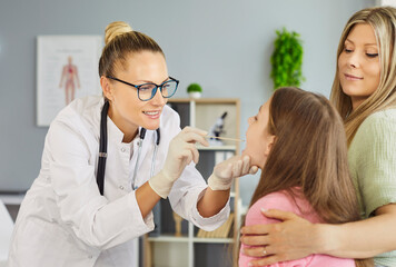 Pediatrician in clinic swabs throat of young girl for medical test while mother offers comfort. Doctor in gloves smiles during procedure. Concept of child healthcare diagnosis and family support.