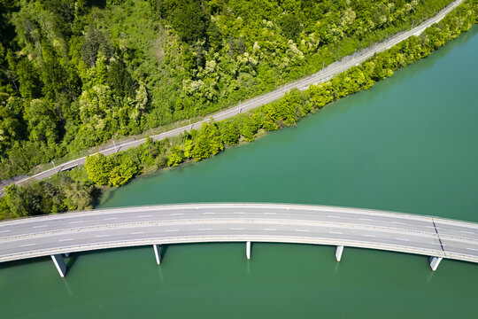 Aerial view of a bridge spanning the vibrant turquoise Drava River, framed by a verdant forest and a narrow road, Ferlach, Carinthia, Austria.