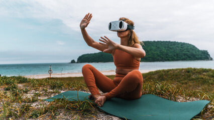 Modern yoga session with virtual twist. Woman choosing suitable workout and practicing breathwork with VR headset near ocean, surrounded by natural beauty.